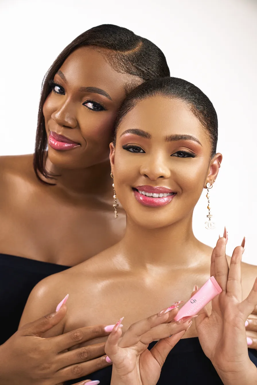 Two smiling models with softly glossed lips, one holding a pink Chardonnay Beauty tube